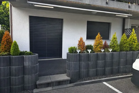 Eingangsbereich eines Gebäudes mit Treppe, Pflanztrögen und Sträuchern.